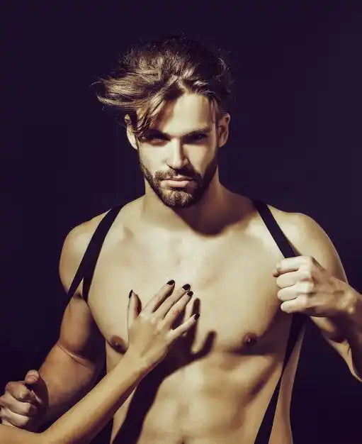 Un homme sans chemise avec une barbe et des bretelles, avec la main d'une femme sur sa poitrine.