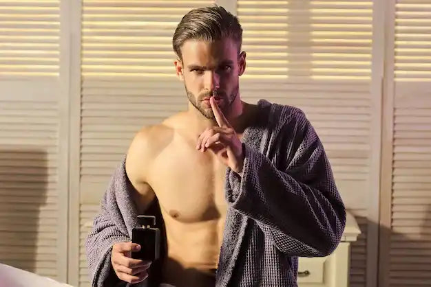 Un homme torse nu portant une robe de chambre grise et tenant une bouteille de parfum pour hommes.